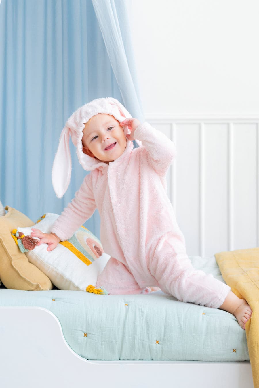 A toddler in a fuzzy pink bunny suit kneeling on a made-up toddler bed, laughing.